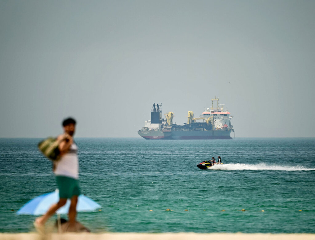 Vorne ein Tourist am Strand von Dubai, hinten Schiffe an der Straße von Hormus: Dreh- und Angelpunkt des Krieges. Foto: AFP